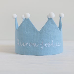May include: A light blue linen crown with white pom poms on top. The crown has the name "Hierom Joshua" embroidered on the front in white thread.
