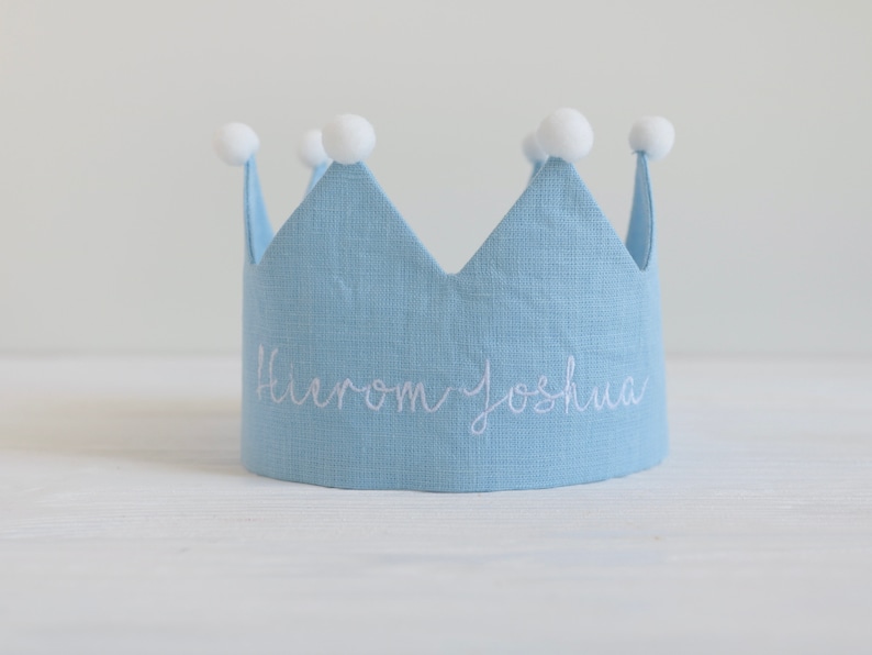 May include: A light blue linen crown with white pom poms on top. The crown has the name "Hierom Joshua" embroidered on the front in white thread.