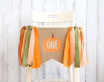 May include: A burlap banner with a pumpkin graphic and the word "ONE" in white, hanging on a white chair. The banner is decorated with orange, green, and tan ribbons, creating a festive look for a first birthday celebration.
