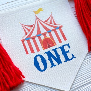 May include: A white fabric banner with a red and white striped circus tent and the word "ONE" in blue. The banner has red tassels on each end.