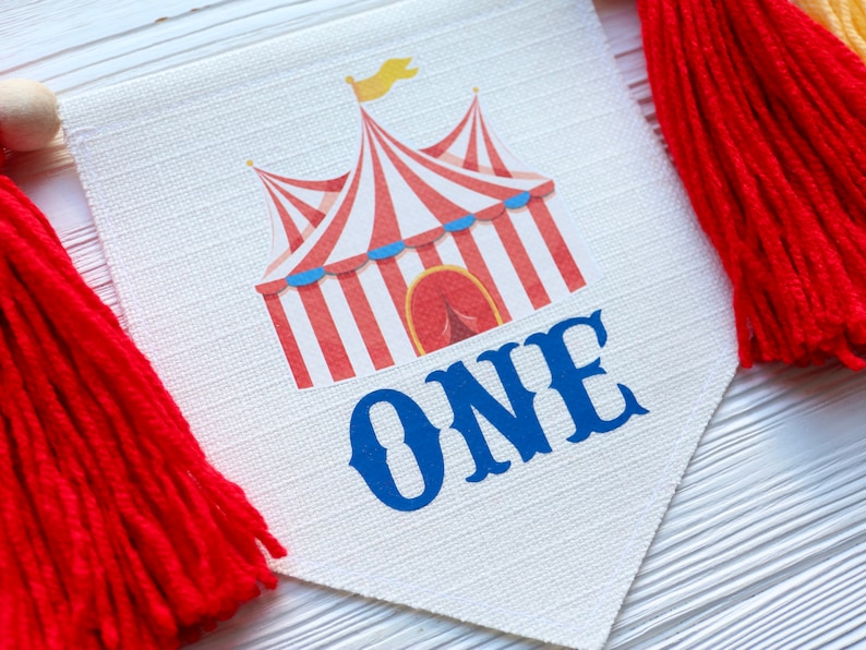 May include: A white fabric banner with a red and white striped circus tent and the word "ONE" in blue. The banner has red tassels on each end.