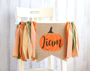 May include: A burlap banner with a pumpkin design and the name "Liam" in brown text. The banner is decorated with orange, green and brown ribbons.