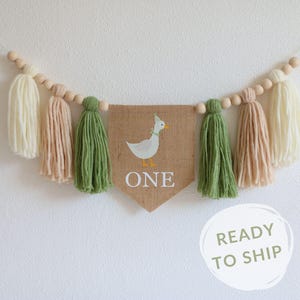 May include: A burlap banner with a white duck illustration and the word "ONE" in white text. The banner is strung with wooden beads and green, cream, and beige yarn tassels.