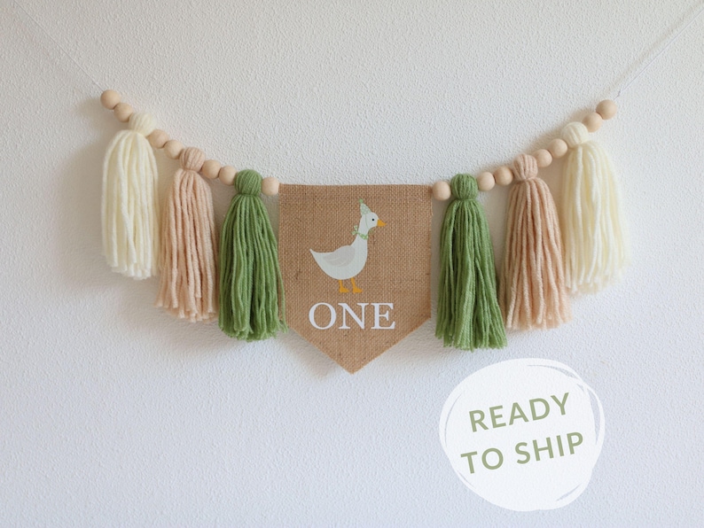 May include: A burlap banner with a white duck illustration and the word "ONE" in white text. The banner is strung with wooden beads and green, cream, and beige yarn tassels.