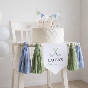 May include: A white cake with a wooden "one" cake topper and a banner that says "Caleb's hole in one" with golf clubs. The cake sits on a white chair decorated with blue, green, and white tassels and wooden beads. Balloons are in the background.