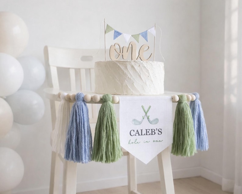 May include: A white cake with a wooden "one" cake topper and a banner that says "Caleb's hole in one" with golf clubs. The cake sits on a white chair decorated with blue, green, and white tassels and wooden beads. Balloons are in the background.