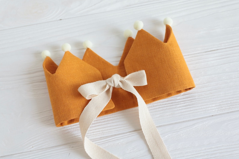 May include: A yellow felt crown with white pom-poms and a white satin ribbon tied in a bow.