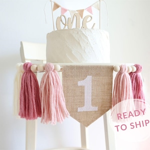 May include: A burlap banner with the number "1" in white glitter, decorated with pink and white yarn tassels. The banner is strung on a wooden dowel with white beads. The banner is ideal for a first birthday party.