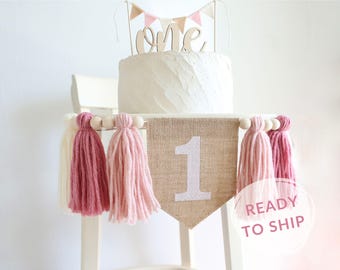 May include: A burlap banner with the number "1" in white glitter, decorated with pink and white yarn tassels. The banner is strung on a wooden dowel with white beads. The banner is perfect for a first birthday party.