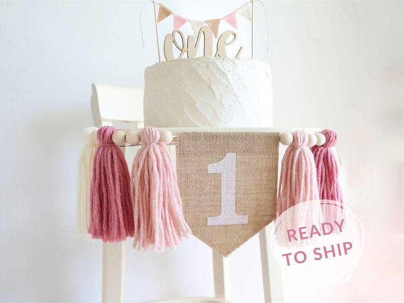 May include: A burlap banner with the number "1" in white glitter, decorated with pink and white yarn tassels. The banner is strung on a wooden dowel with white beads. The banner is ideal for a first birthday party.
