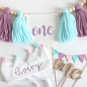 May include: A white felt crown with the word "every" embroidered in purple, decorated with colorful pom-poms. A white banner with the word "one" in purple script hangs above a wooden cake topper with the word "one" in cursive.