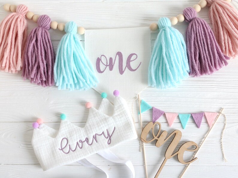 May include: A white felt crown with the word "every" embroidered in purple, decorated with colorful pom-poms. A white banner with the word "one" in purple script hangs above a wooden cake topper with the word "one" in cursive.