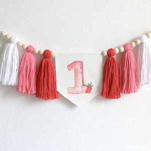 Puede incluir: Una guirnalda de borlas rosa y blanca con una pancarta blanca que presenta el n&uacute;mero "1" decorado con acuarela rosa y una fresa.