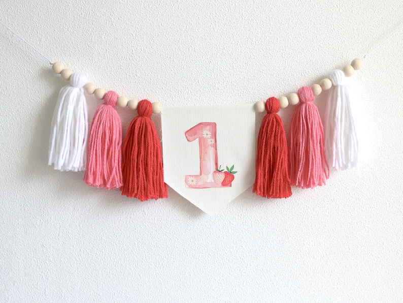 Puede incluir: Una guirnalda de borlas rosa y blanca con una pancarta blanca que presenta el n&uacute;mero "1" decorado con acuarela rosa y una fresa.