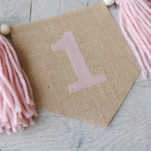 May include: A burlap banner with the number "1" in pink, decorated with pink and burgundy yarn tassels.