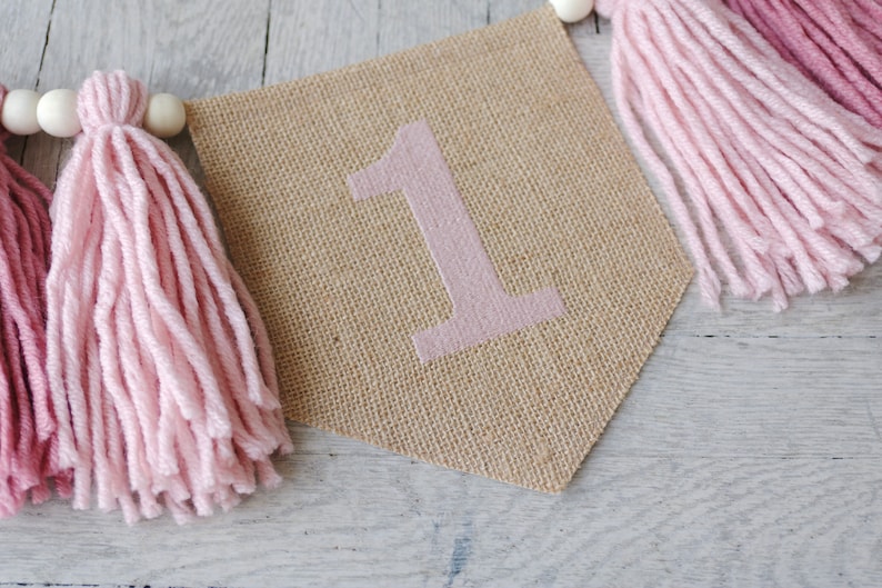 May include: A burlap banner with the number "1" in pink, decorated with pink and burgundy yarn tassels.