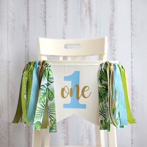 Puede incluir: Una silla alta blanca con una pancarta de tela azul y verde que dice "one" en letras doradas. La pancarta tiene cintas verdes, azules y marrones colgando.