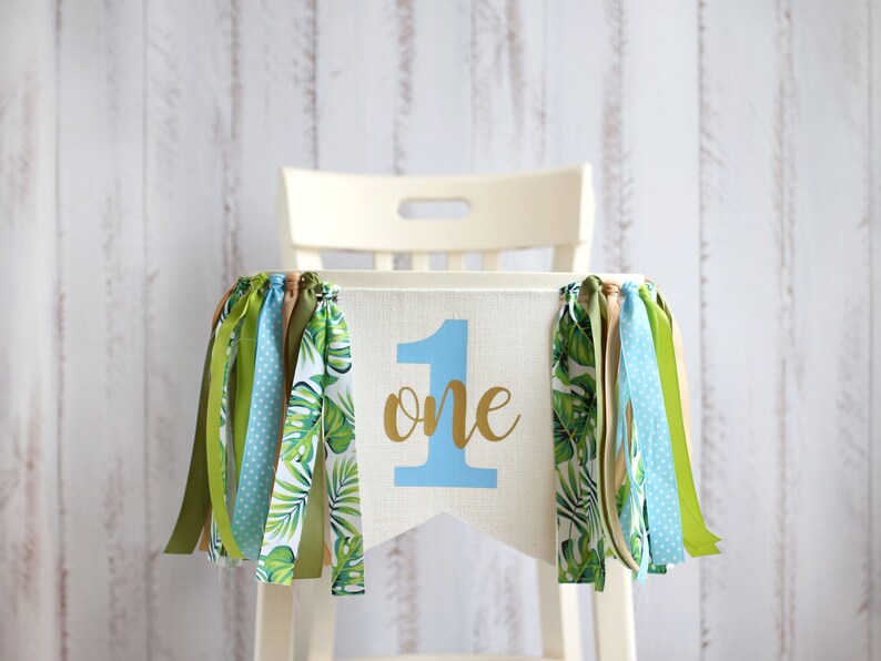 Puede incluir: Una silla alta blanca con una pancarta de tela azul y verde que dice "one" en letras doradas. La pancarta tiene cintas verdes, azules y marrones colgando.