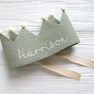 May include: A green linen crown with white pom poms and the name "Harrison" embroidered on it. The crown has a white ribbon attached.