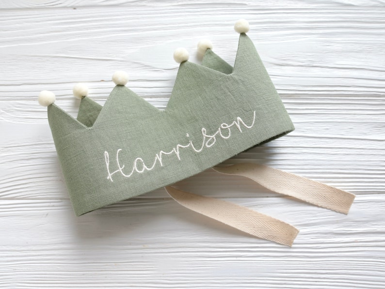 May include: A green linen crown with white pom poms and the name "Harrison" embroidered on it. The crown has a white ribbon attached.
