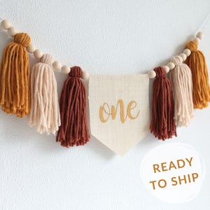 May include: A burlap banner with the word "one" embroidered in gold thread, strung with wooden beads and yarn tassels in shades of mustard yellow, cream, and burgundy.