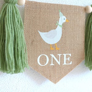 May include: A burlap banner with a green tassel on each end. The banner features a white goose with a yellow beak and a green hat. The banner also has the word "ONE" printed in white.