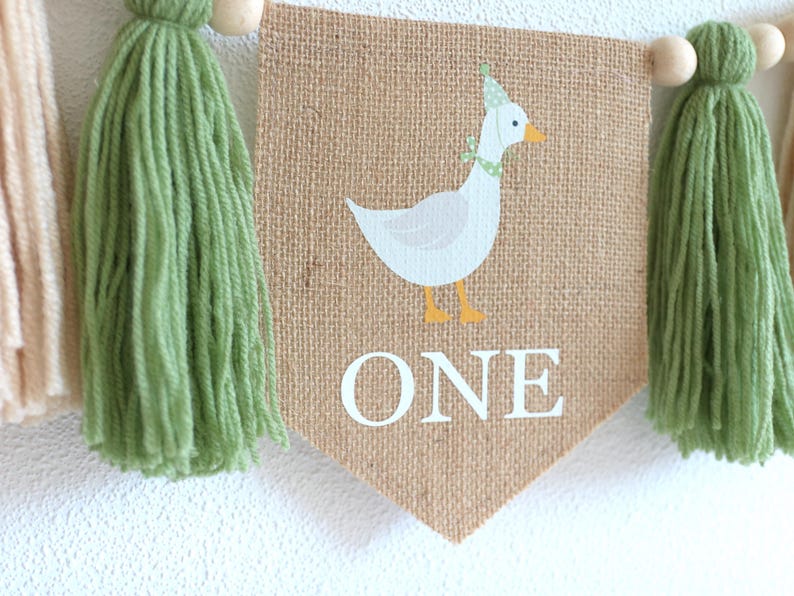 May include: A burlap banner with a green tassel on each end. The banner features a white goose with a yellow beak and a green hat. The banner also has the word "ONE" printed in white.