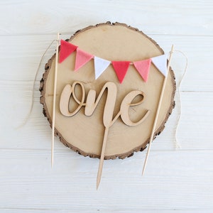 Puede incluir: Topper de pastel de madera con la palabra "one" en escritura cursiva, decorado con una guirnalda de fieltro rosa y blanco.