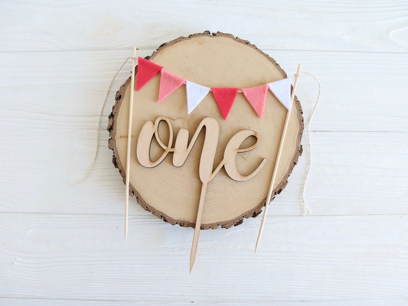 Puede incluir: Topper de pastel de madera con la palabra "one" en escritura cursiva, decorado con una guirnalda de fieltro rosa y blanco.