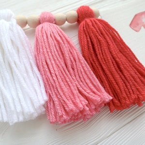 Puede incluir: Tres borlas de hilo en blanco, rosa y rojo colgando de una varilla de madera. Las borlas est&aacute;n hechas de hilo suave y esponjoso y son perfectas para agregar un toque de fantas&iacute;a a cualquier espacio.
