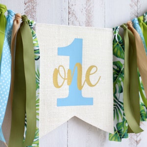 Puede incluir: Una pancarta blanca con el n&uacute;mero "1" en azul y la palabra "one" en dorado. La pancarta est&aacute; decorada con cintas verdes y azules.
