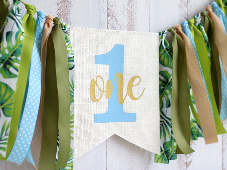 Puede incluir: Una pancarta blanca con el n&uacute;mero "1" en azul y la palabra "one" en dorado. La pancarta est&aacute; decorada con cintas verdes y azules.