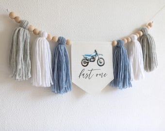Puede incluir: Una guirnalda de borlas azules y blancas con una pancarta blanca que dice "fast one" y una ilustración de acuarela de una moto de cross.