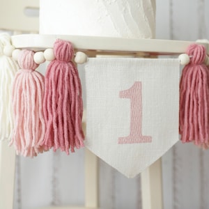 May include: A white high chair with a pink and white tassel garland and a white banner with the number "1" in pink.