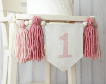May include: A white high chair with a pink and white tassel garland and a white banner with the number "1" in pink.