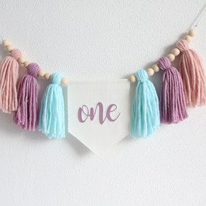 May include: A white banner with the word "one" written in purple script hangs from a string of wooden beads with pink, purple, and light blue yarn tassels.