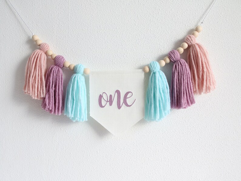May include: A white banner with the word "one" written in purple script hangs from a string of wooden beads with pink, purple, and light blue yarn tassels.
