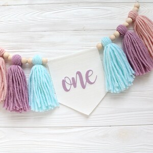 May include: A white banner with the word "one" written in purple script hangs from a string of wooden beads with pink, purple, and blue yarn tassels.