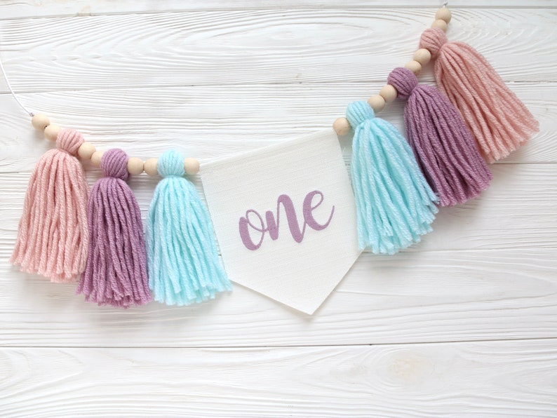 May include: A white banner with the word "one" written in purple script hangs from a string of wooden beads with pink, purple, and blue yarn tassels.