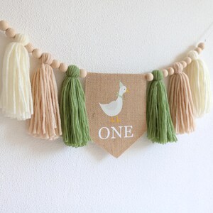 May include: A burlap banner with a white duck and the word "ONE" in white letters. The banner is strung with wooden beads and green, cream, and beige yarn tassels.