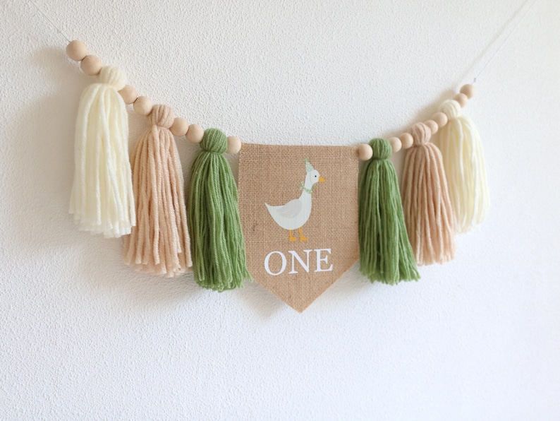 May include: A burlap banner with a white duck and the word "ONE" in white letters. The banner is strung with wooden beads and green, cream, and beige yarn tassels.