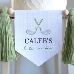 May include: A banner with the text "CALEB'S hole in one" and a watercolor illustration of crossed golf clubs. The banner is white with green text and accents. It is decorated with green and blue tassels and wooden beads.