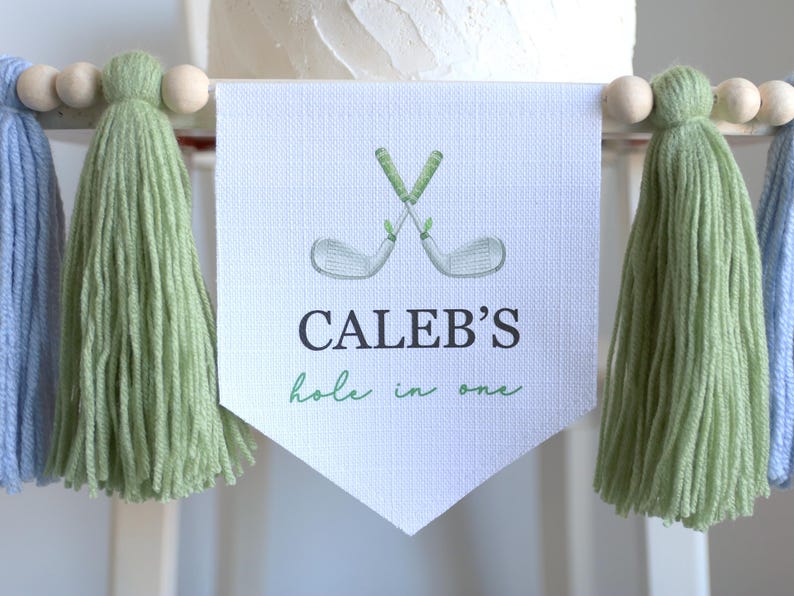 May include: A banner with the text "CALEB'S hole in one" and a watercolor illustration of crossed golf clubs. The banner is white with green text and accents. It is decorated with green and blue tassels and wooden beads.