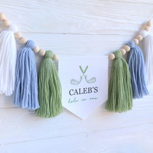 May include: A decorative banner with white, blue, and green tassels and wooden beads. The banner features a white pennant with the text "CALEB'S hole in one" and a golf club graphic. The banner is designed for a golf-themed event.