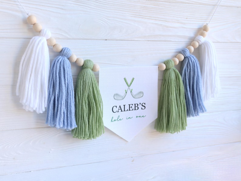 May include: A decorative banner with white, blue, and green tassels and wooden beads. The banner features a white pennant with the text "CALEB'S hole in one" and a golf club graphic. The banner is designed for a golf-themed event.