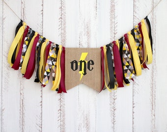 May include: A burlap banner with the word "one" in black lettering and a yellow lightning bolt. The banner is decorated with yellow, black, and burgundy ribbon streamers.