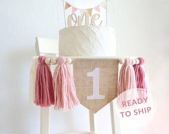 May include: A burlap banner with the number "1" in white glitter, decorated with pink and white yarn tassels. The banner is strung on a wooden dowel with white beads. The banner is perfect for a first birthday party.
