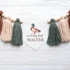 以下が含まれることがあります： 緑色のアヒルのイラストと「one lucky duck WALTER」というテキストが書かれた白いバナー。バナーは茶色、ベージュ、緑の糸のタッセルで飾られています。