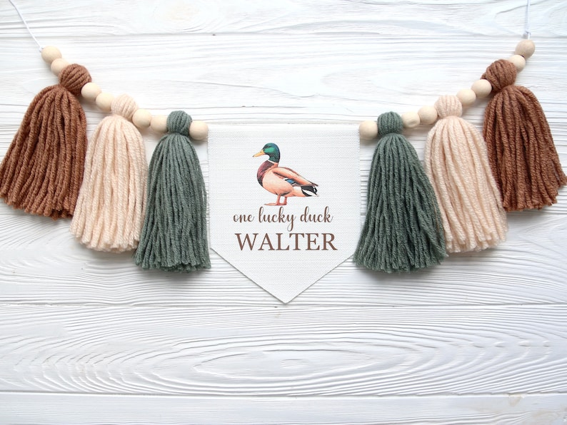 以下が含まれることがあります： 緑色のアヒルのイラストと「one lucky duck WALTER」というテキストが書かれた白いバナー。バナーは茶色、ベージュ、緑の糸のタッセルで飾られています。