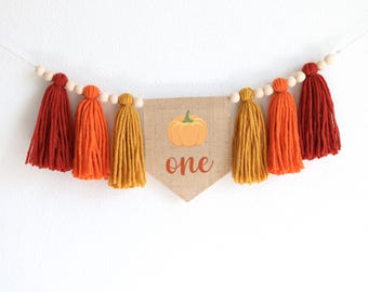 May include: A decorative banner with a burlap pennant displaying a pumpkin illustration and the word "one". The banner is decorated with orange, yellow, and red tassels, and wooden beads. Ideal for a first birthday party.